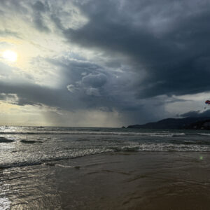 Parasail over Patong Beach beneath storm clouds with sunbeams and wet-sand reflections, Phuket, Thailand.