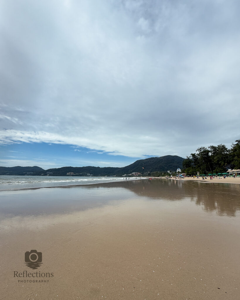 Calm reflective shoreline at Patong Beach, Phuket, showing wet sand mirroring soft blue sky under light cloud cover.