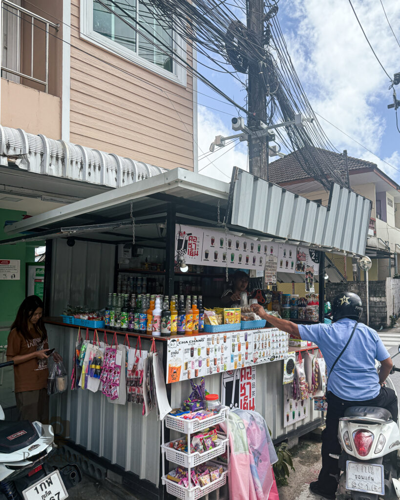 Street vendor serving a motorbike rider in Patong, Phuket, with power lines and local buildings in the background.