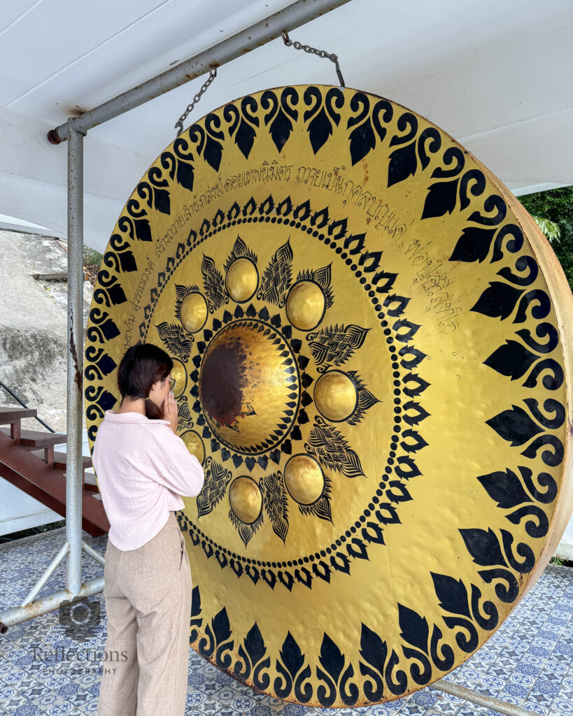 A woman prays before a large golden temple gong with black decorative patterns at Wat Khao Rang Samakkhitham in Phuket, Thailand.