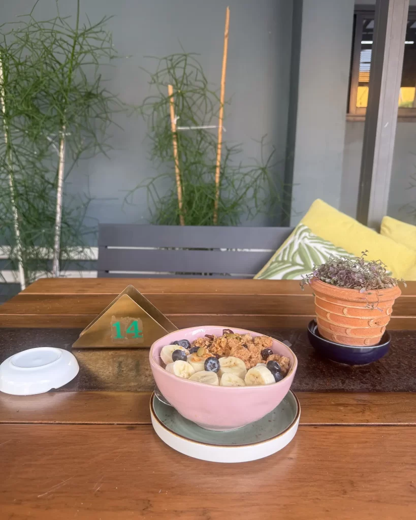 Wide photo of a breakfast bowl on a café table with extra objects and background clutter, making the subject feel unclear.