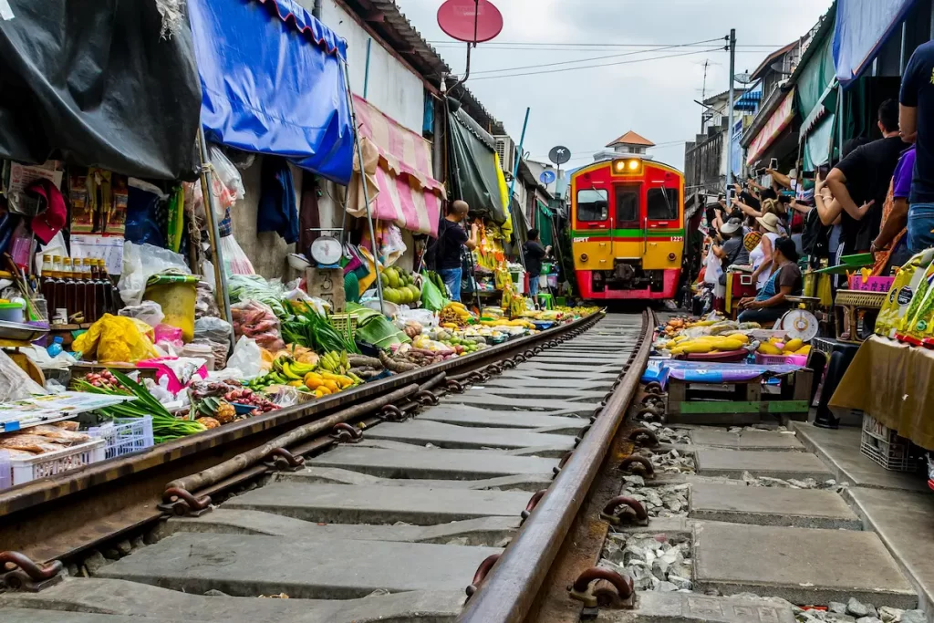 Train passing through the Maeklong Railway Market in Thailand with vendors and fresh produce along the tracks.