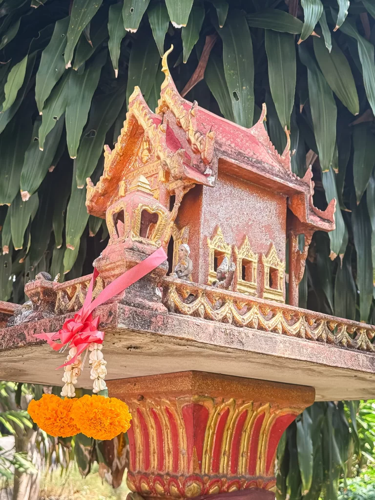 Weathered Thai spirit house with marigold garlands and offerings in Patong, Phuket, photographed in natural light.